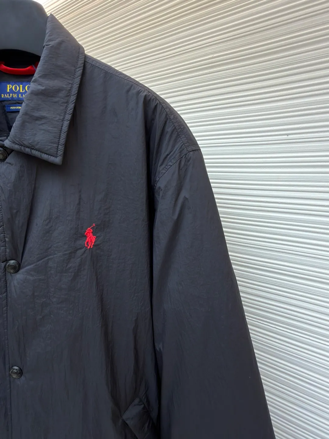 Ralph Lauren red label light cotton jacket (coach jacket) ●Original version: purchased and developed for 2699 ●Item number: 1681910-B90 ●Fabric: high-density nylon taffeta fabric ●Technology: embroidery production ●Color: black ●Version: fitted version ●Weight: about 340 grams ●Size: S-M-L-XL ●Description: The density of high-density nylon taffeta is about 210T-300T, with a smooth and slightly matte surface, excellent windproof and waterproof properties (with DWR water-repellent treatment), strong wrinkle resistance and easy care, in line with the positioning of American casual "durable and practical". Classic dark black, using vat dyeing process (dye penetrates into the fiber), the color fastness reaches ISO level 4 or above, preventing fading or color transfer after washing. The lightweight cotton-polyester blend lining (65% Polyester + 35% Cotton) reduces the friction between the fabric and inner clothing, improving the smoothness of wearing; the blended material combines breathability and durability to avoid the stuffy feeling of pure nylon lining. The classic Coach Jacket has a loose fit with slightly dropped shoulder lines (shoulder width is 1-2cm wider than the standard fit). The length reaches the hips and the sleeves are slightly tapered straight sleeves, taking into account both freedom of movement and casual style. Polo horse logo on the left chest: flat satin stitch embroidery (satin embroidery), embroidery thread: 100% polyester high color fastness embroidery thread (the color number is consistent with the brand's standard red color card), stitch density 12-15 stitches/cm, the outline of the horse logo is clear, and the details of the rider and horse are burr-free. Embroidery positioning: precise positioning through the template (12cm from the neckline and 8cm from the placket) to ensure left and right symmetry and centering. Topstitching technology: The seams of the body are made of double-needle flat seams (needle pitch 3cm/10 needles), the stitches are smooth and straight, the stitching color is dark black (the same color as the fabric), and the stitch width is 0.5cm, which enhances firmness while forming simple decorative lines. The splicing of the front placket and the body: the placket is made of double-layer folding + flat seam splicing, with a thickness of about 0.3cm to ensure that the buttons are not easily deformed when fixed; the edges of the placket are cut with a hot knife to avoid burrs. The sleeves are spliced ​​to the body: the armholes are spliced ​​with round-head keyhole machine seams + flat seams. After splicing, the armholes have a natural curvature and no sense of tightness during movement; the height of the sleeves is about 8-10cm, which is in line with the loose characteristics of casual jackets. Main button: black urea resin button, matte surface treatment, buttonholes are hand-made keyholes (6 strands of cotton thread, keyhole density 8 stitches/mm), and the edges of the eyelets are smooth and without burrs. Collar buttons: The first button on the collar is a hidden button (diameter 12mm), which prevents the collar from opening and improves the neatness of wearing.